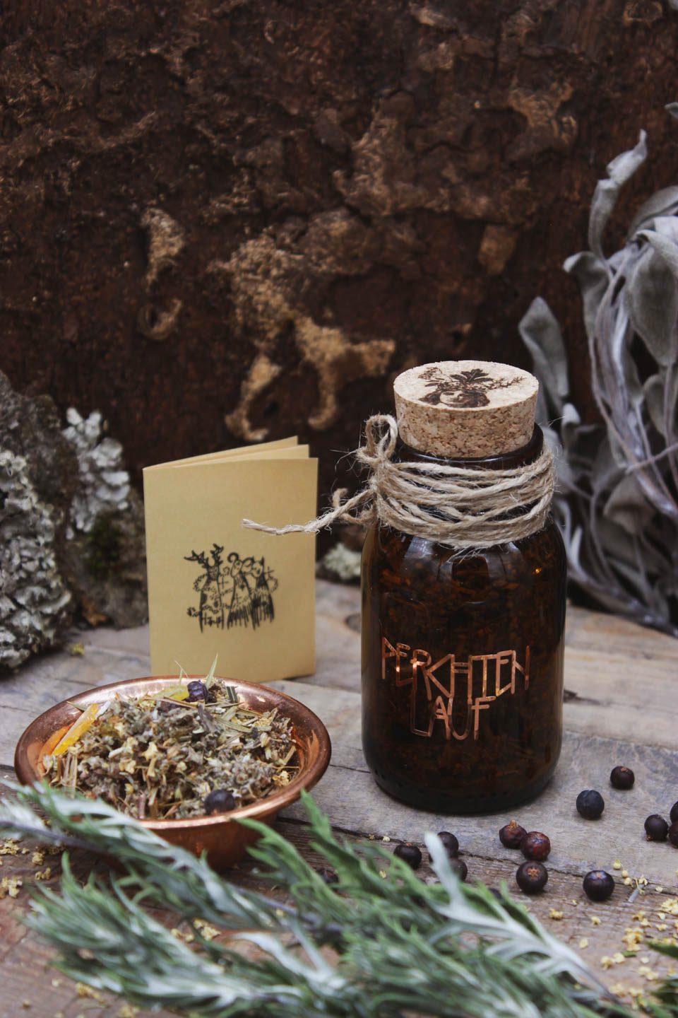 Räucherwerk in einem Braunglas mit Korken. Darauf steht in kupferner Schrift Perchtenlauf. Daneben steht eine Kupferschale gefüllt mit Räucherwerk. Drumherum liegen Wacholderbeeren verstreut. Im Hintergrund ist ein gefalteter Zettel zu sehen auf denen verkleidete Gestalten, die Perchten, aufgedruckt sind.