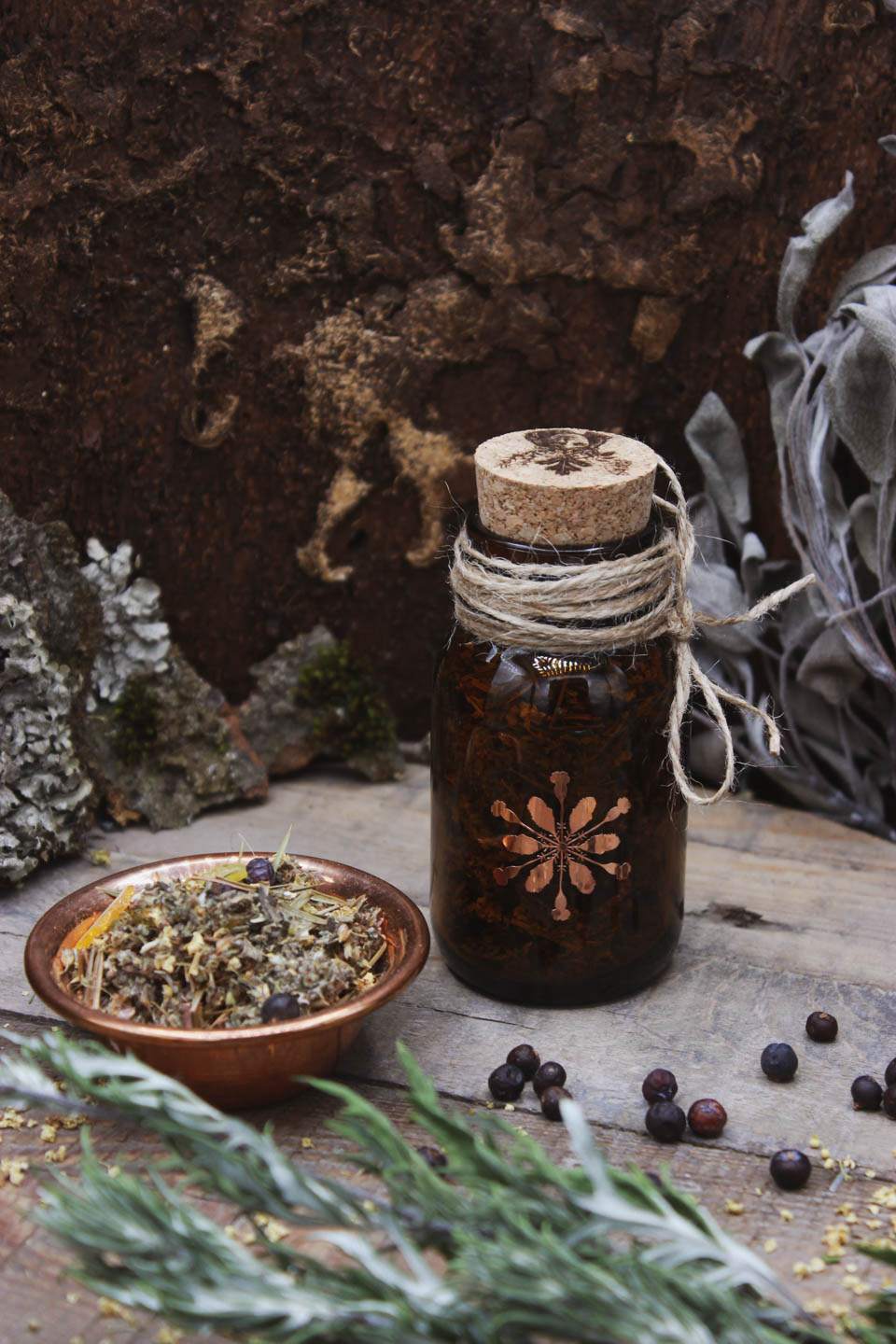 Räucherwerk in einem Braunglas mit Korken. Darauf ist in kupferfarben das Hollenkraut-Logo zu sehen. Daneben steht eine Kupferschale gefüllt mit Räucherwerk. Drumherum liegen Wacholderbeeren verstreut.