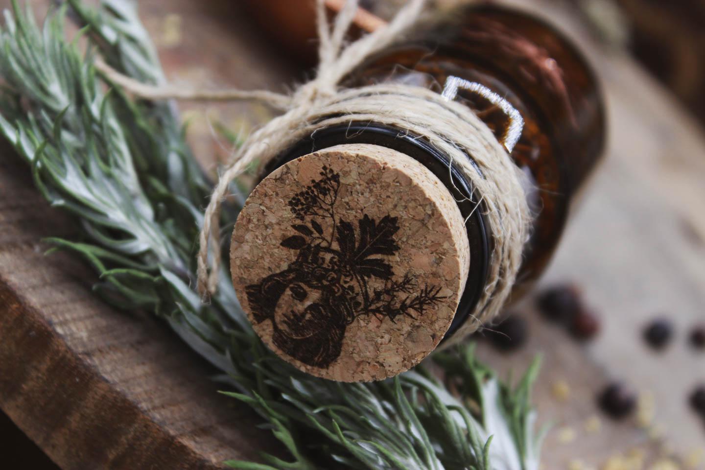 Nahaufnahme des Korkens von oben. Auf den Korken ist ein Motiv eingebrannt. Es zeigt die Perchta mit einer Gesichtshälfte im Schatten. Sie trägt eine Krone aus Holunder, Wacholder und Beifuß.