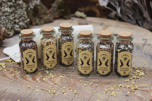 Sechs Glasflaschen mit Korken, gefüllt mit getrockneten Kräutern, stehen auf einem rustikalen Holztisch. Auf den Etiketten steht von links nach rechts: Maiennacht, Taumond, Perchtenlauf, Bärenruhe, Trotula und Pech & Gold.