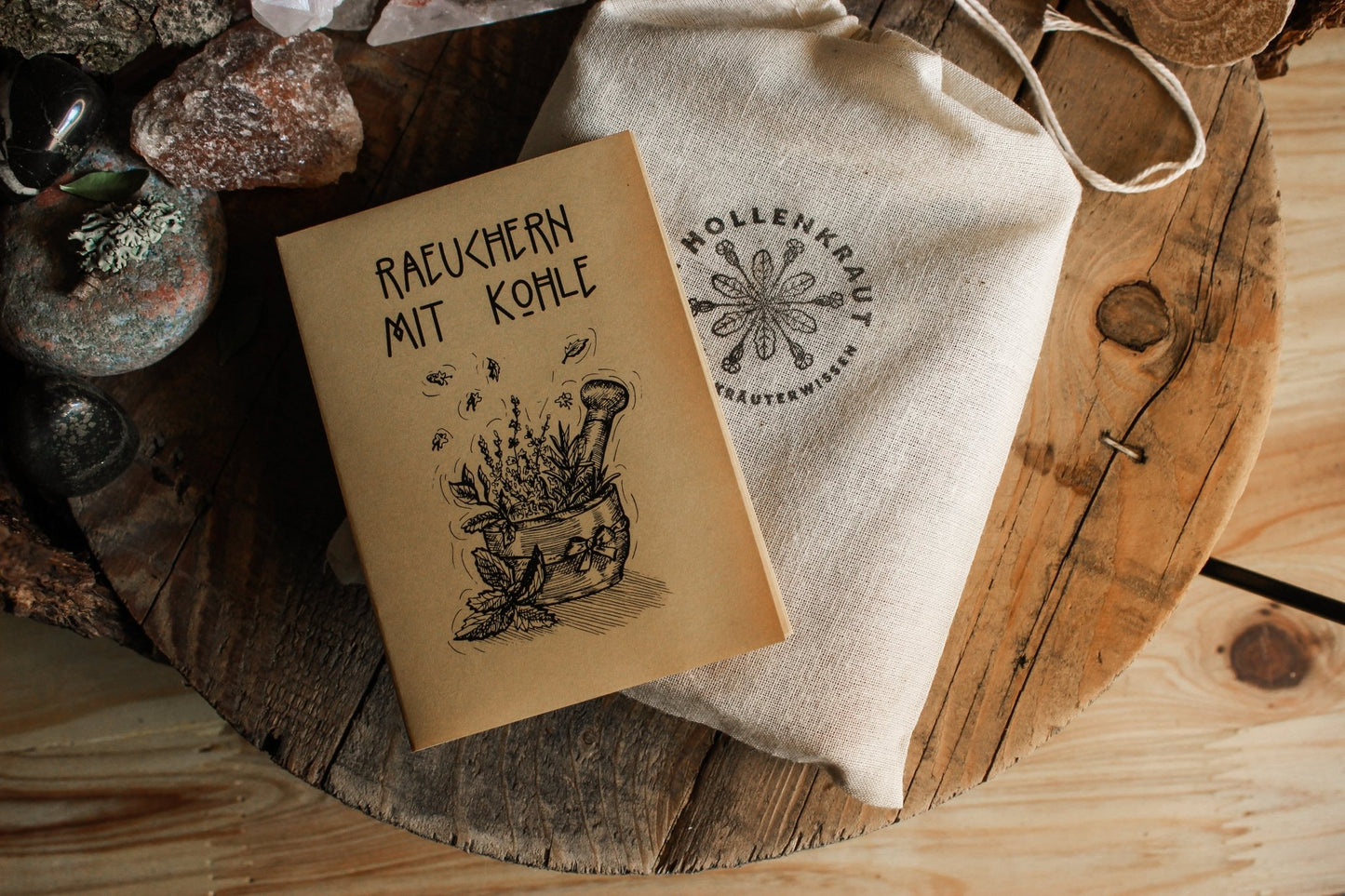 Rustikale Szene mit einem Kräuter-Booklet „Räuchern mit Kohle“ und einem naturfarbenen Stoffbeutel „Hollenkraut“ auf Holzuntergrund. Umgeben von Kristallen, Steinen und getrockneten Pflanzen.
