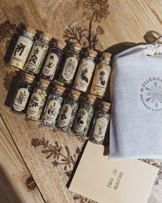 12 Räucherungen, Edition I - 12 Räucherungen, Edition I - Sammlung von zwölf Kräuterflaschen, darunter Fichtennadeln, Wacholder und Salbei, präsentiert auf einem hölzernen Schneidebrett mit floralen Gravuren. daneben ein kleines Booklet und eine kleine Leinentasche