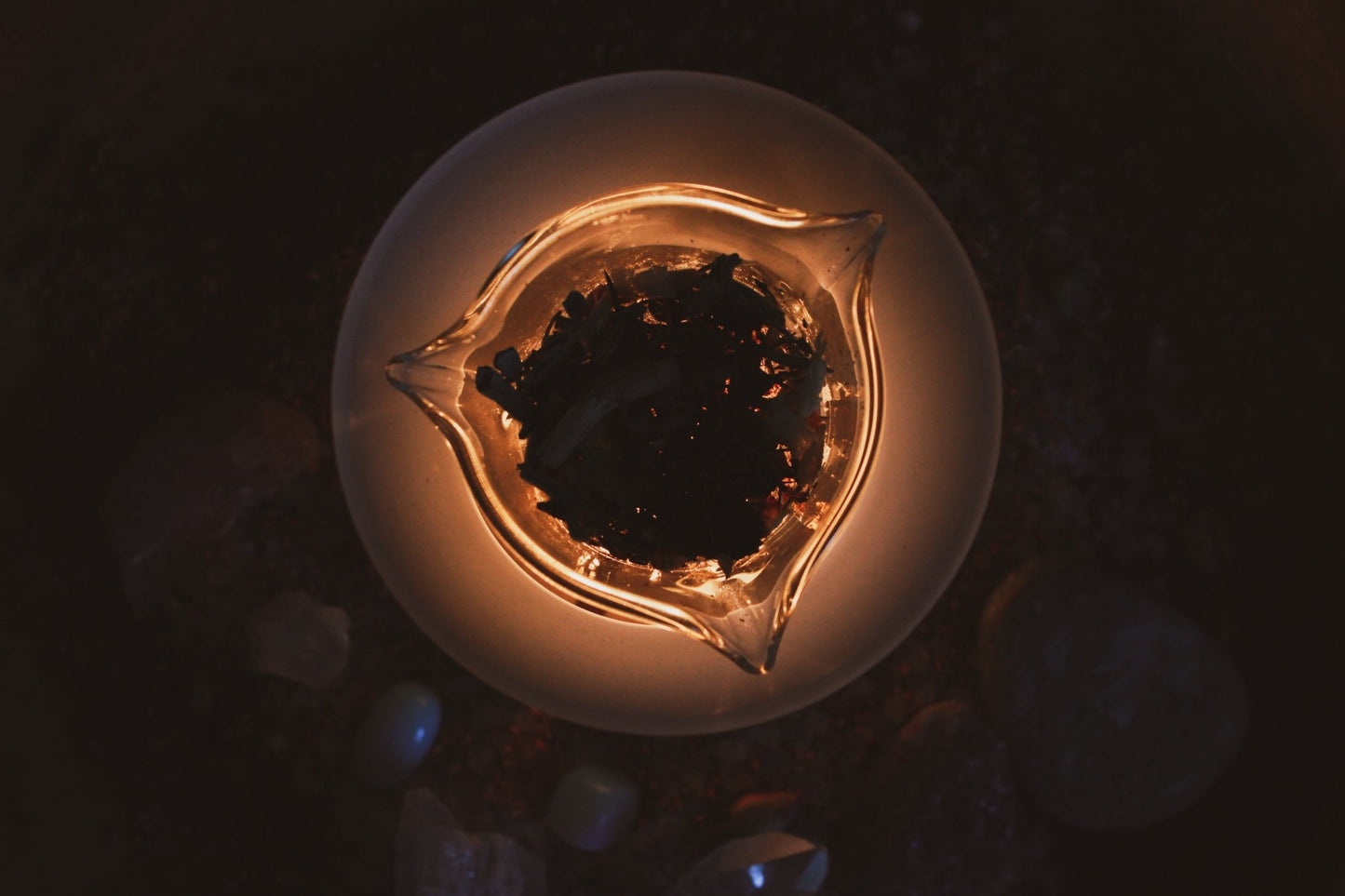 Foto von oben, Duftlampe für Räuchermischungen und lose Kräuter, Räucherung im Glasbehälter, umgeben von einer Auswahl an spirituellen Heilkristallen und Edelsteinen, in einer atmosphärischen, warm beleuchteten Umgebung