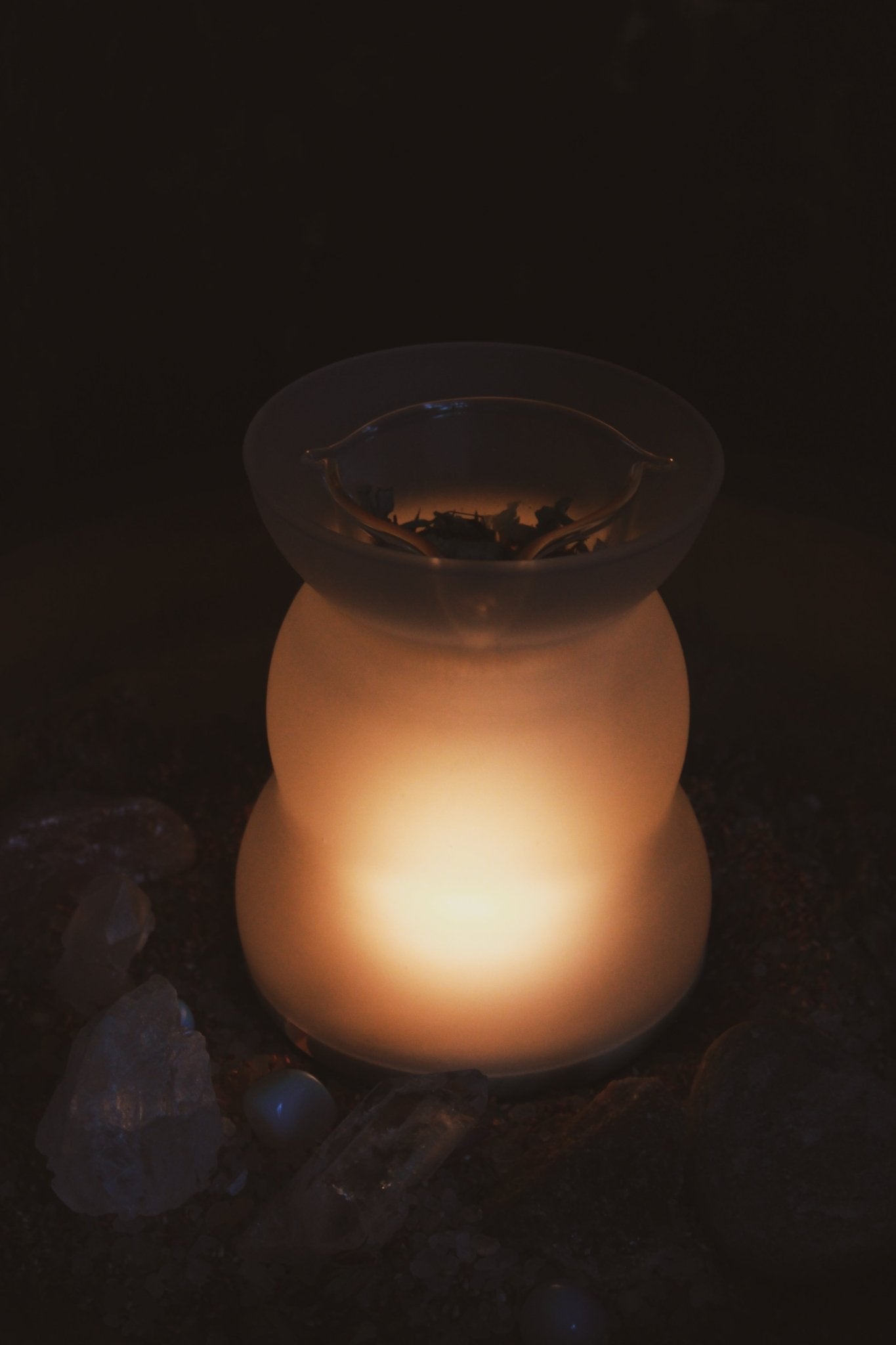Angezündete Duftlampe für Räuchermischungen und lose Kräuter - Elegante, graue gläserne Duftlampe, präsentiert vor einer rustikalen Holzwand, begleitet von einem klaren Glaseinsatz mit komplizierten Designs und einem hölzernen Sockel. Es ist dunkel und nur das warme Licht hinter dem milchigen Glas ist klar zu sehen