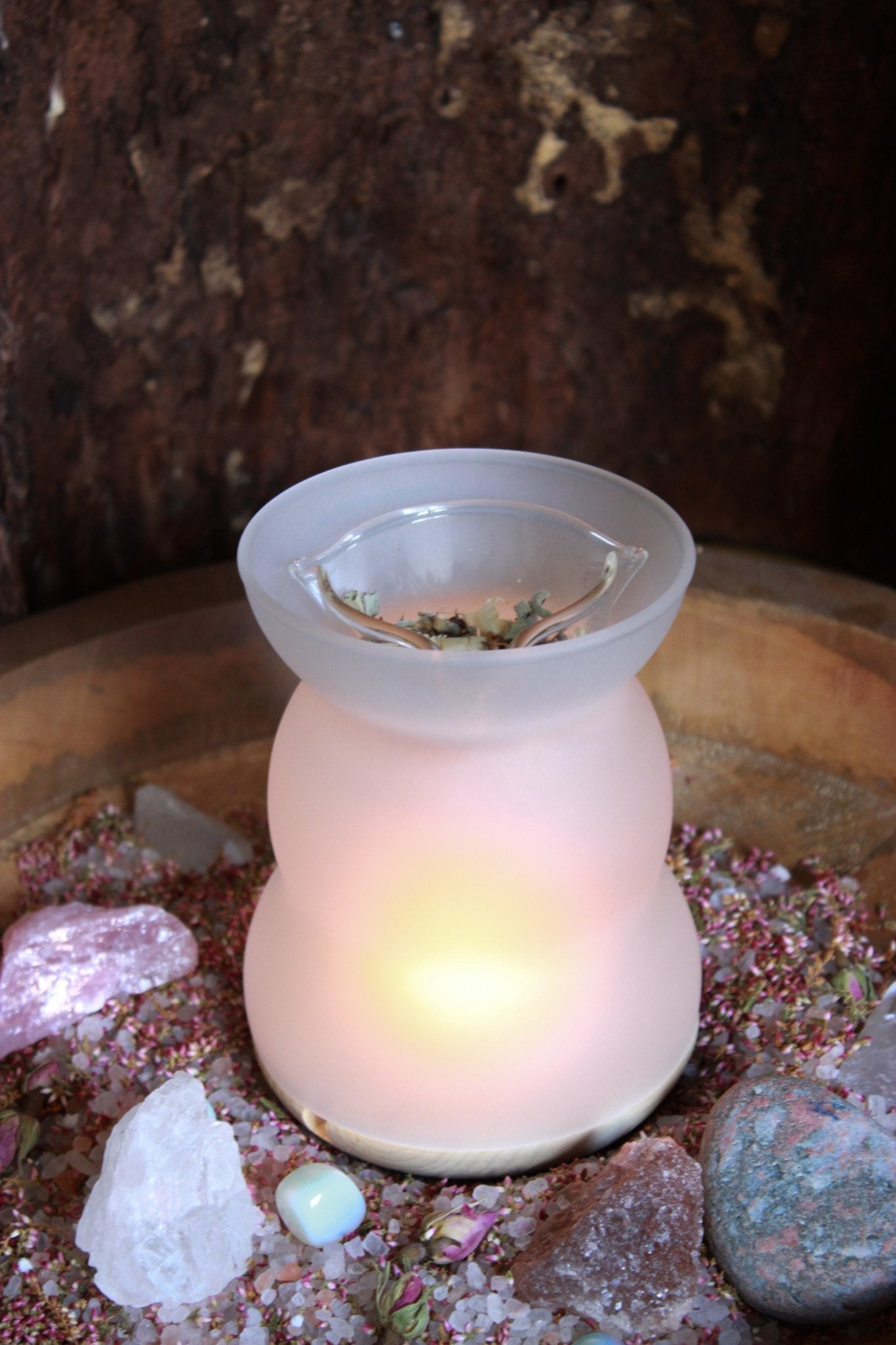 Duftlampe für Räuchermischungen und lose Kräuter - Moderne, weiße Duftlampe mit einer klaren Schale oben, gefüllt mit getrockneten Blättern, präsentiert auf einem Holztisch, umgeben von einer Auswahl an bunten Kristallen und getrockneten Blumen. 