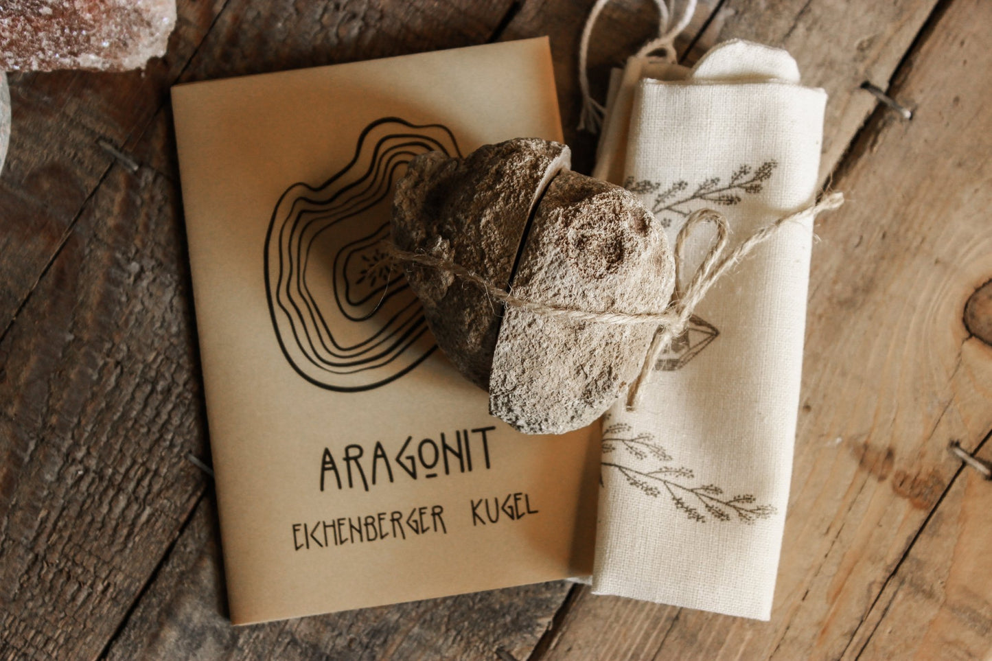 Das Bild zeigt eine in zwei Hälften geschnittene Eichenberger Kugel, die mit einer Schnur zusammen gebunden ist. Unter dem Stein liegt ein Jutebeutel und ein beige-farbenes Zine mit der Aufschrift: Aragonit Eichenberger Kugel