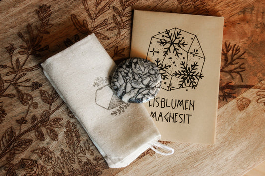 Eisblumen-Magnesit Scheibenstein aus Österreich - Faire regionale Steine Handgefertigtes Produktset bestehend aus einer bedruckten Leinentasche, einem Marmor-Magnesit und einem illustrierten Booklet mit dem Text ‘EISBLUMEN MAGNESIT’, präsentiert auf einem kunstvoll verzierten Holztisch.