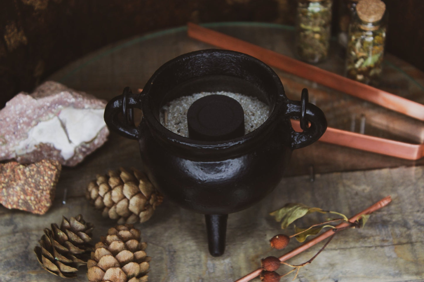 Kohle in einem schwarzem Kessel, umgeben von natürlichen Elementen und Kräutern, eine Kupferzange liegt daneben