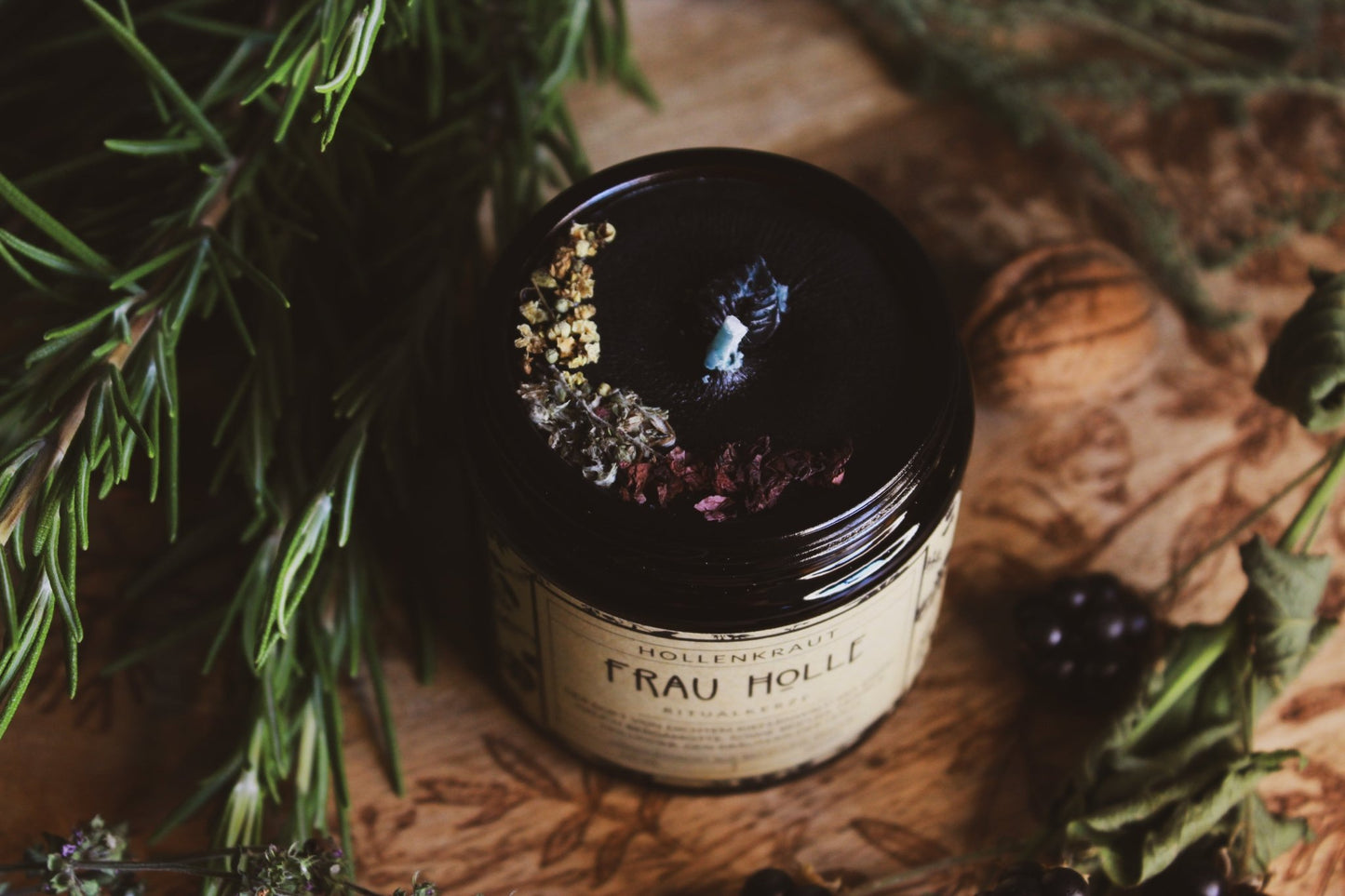Ein sorgfältig gestaltetes Bild zeigt ein Glas ‘Frau Holle Ritualkerze’ von oben, umgeben von natürlichen Elementen wie Beeren, Kräutern und einer Walnuss auf einem hölzernen Hintergrund. Die Kerze, verpackt in einem etikettierten Glas, strahlt eine Atmosphäre der Ruhe und Entspannung aus. Der Duft von dünnem Kiefernwald mit einem Hauch Bergamotte und Kiefernnuss, Mohn & Holunder, den sowie der Hölle handgemacht aus Recycling Wachs 120 ml Vegan.