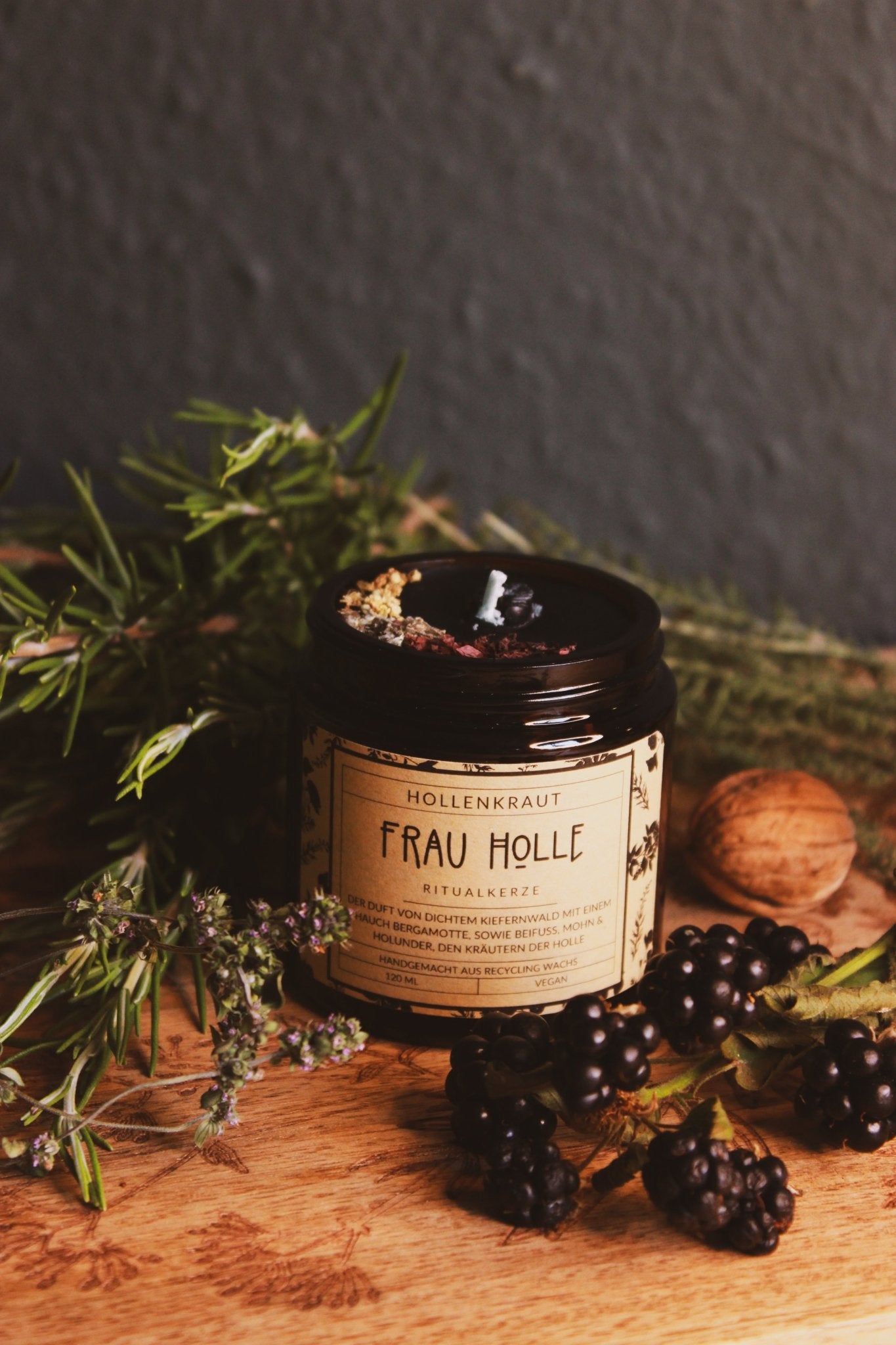 Ein sorgfältig gestaltetes Bild zeigt ein Glas ‘Frau Holle Ritualkerze’, umgeben von natürlichen Elementen wie Beeren, Kräutern und einer Walnuss auf einem hölzernen Hintergrund. Die Kerze, verpackt in einem etikettierten Glas, strahlt eine Atmosphäre der Ruhe und Entspannung aus. Der Duft von dünnem Kiefernwald mit einem Hauch Bergamotte und Kiefernnuss, Mohn & Holunder, den sowie der Hölle handgemacht aus Recycling Wachs 120 ml Vegan.
