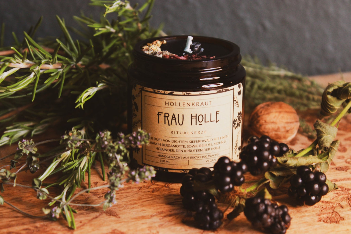 Kräuter-Kerze "Frau Holle" - Handgemachte Frau Holle Ritualkerze aus Recyclingwachs in einem dunklen Glasbehälter, umgeben von natürlichen Elementen wie grünen Kräutern, Brombeeren und einer Walnuss auf einer hölzernen Oberfläche. Die Kerze verströmt einen Duft von dichtem Kiefernwald, ergänzt durch Noten von Beifuß, Mohn und Holunder, den Kräutern der Holle.