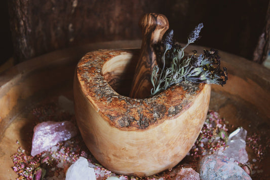 Mörser & Stößel aus Olivenholz - Mörser & Stößel aus Olivenholz - Rustikales, handgefertigtes Mörser- und Stößelset aus natürlichem Holz, gefüllt mit getrockneten Kräutern und umgeben von Heilsteinen und getrockneten Blumen, auf einem dunklen, texturierten Hintergrund.