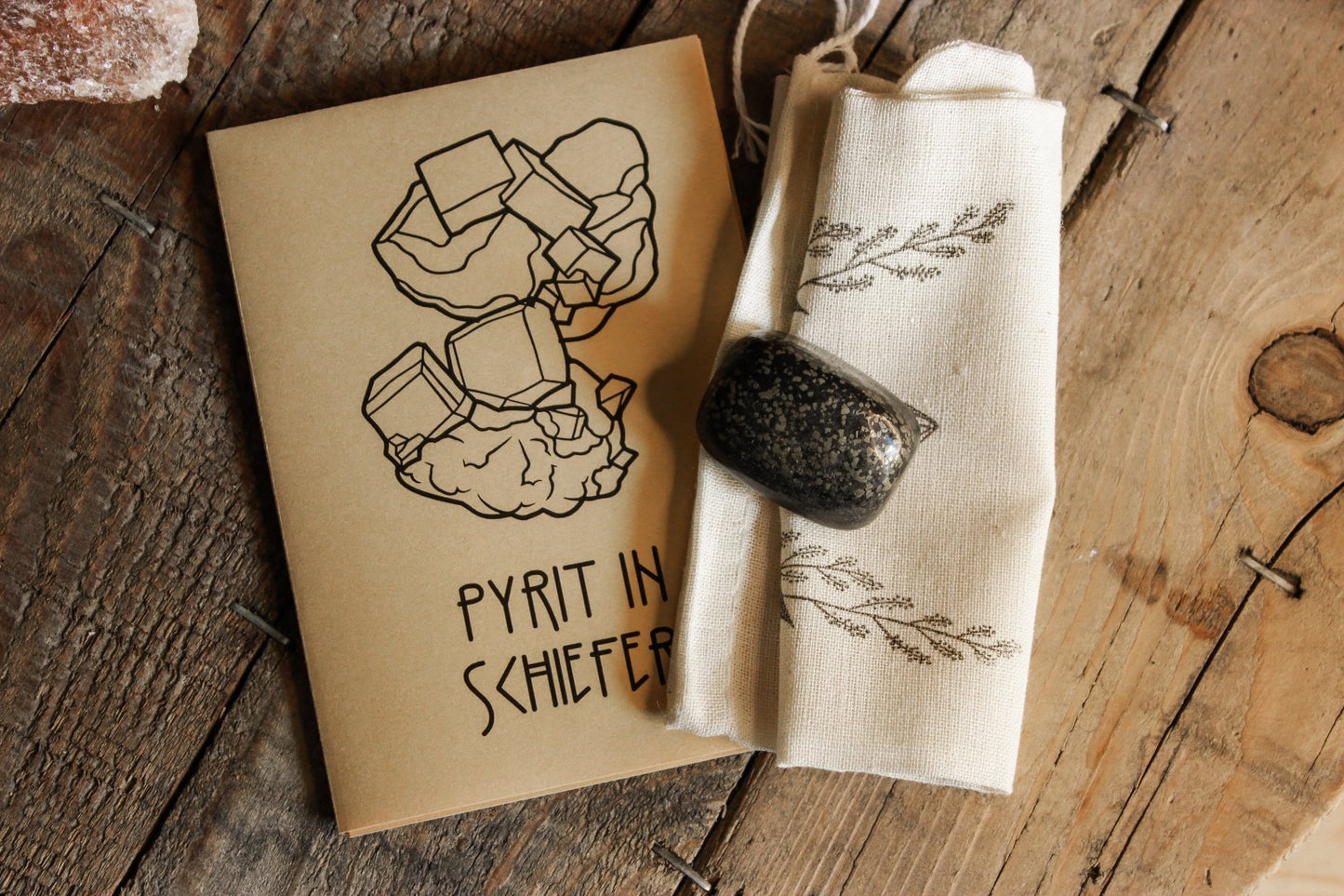 Pyrit in Schiefer Trommelstein aus Deutschland - Zine ‘PYRIT IN SCHIEFER’ neben einem Stück Pyrit und einer Stofftasche mit Blattdesigns, präsentiert auf einem rustikalen Holztisch.