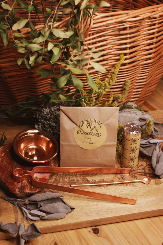 Räucherset: Zange, Schale, Löffel, Räucherkohle und Sand - Ein harmonisches Bild, das ein Paket von ‘RAUCHERSAND 200g 100% Quartz’ auf einem Holzbrett darstellt, neben einem Kupferteller und einem Kupferlöffeln und einer Kupferzange. Ein gerollter Artikel mit komplexen Designs, eingewickelt in klarem Kunststoff, liegt neben dem Paket. Im Hintergrund ist ein geflochtener Korb mit grünen Pflanzen und einigen gelben Blumen zu sehen.