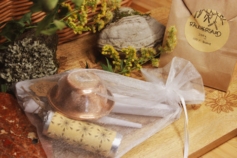 Räucherset: Zange, Schale, Löffel, Räucherkohle und Sand - Ein harmonisches Arrangement von natürlichen und spirituellen Elementen auf einer Holzoberfläche, einschließlich eines Beutels mit ‘RAUCHERSAND 200g 100% Quartz’, einem dekorativen Stein mit komplexen Texturen und Mustern, einer Kerze, und blühenden Pflanzen mit gelben Blüten.