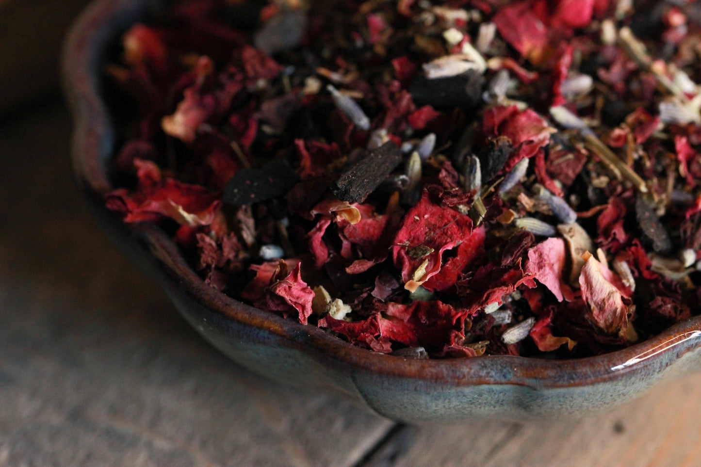 Rustikale Keramikschale, gefüllt mit einer Mischung aus getrockneten Blumen und Kräutern auf einem Holztisch. Die Schale ist prall gefüllt mit roten und rosa Blütenblättern, dunklen Blättern und anderen organischen Elementen. Die Textur der getrockneten Blumen ist deutlich sichtbar; sie wirken knusprig und zart. Die Beleuchtung ist weich und gedämpft, wirft sanfte Schatten und hebt die Texturen der getrockneten Kräutern hervor.