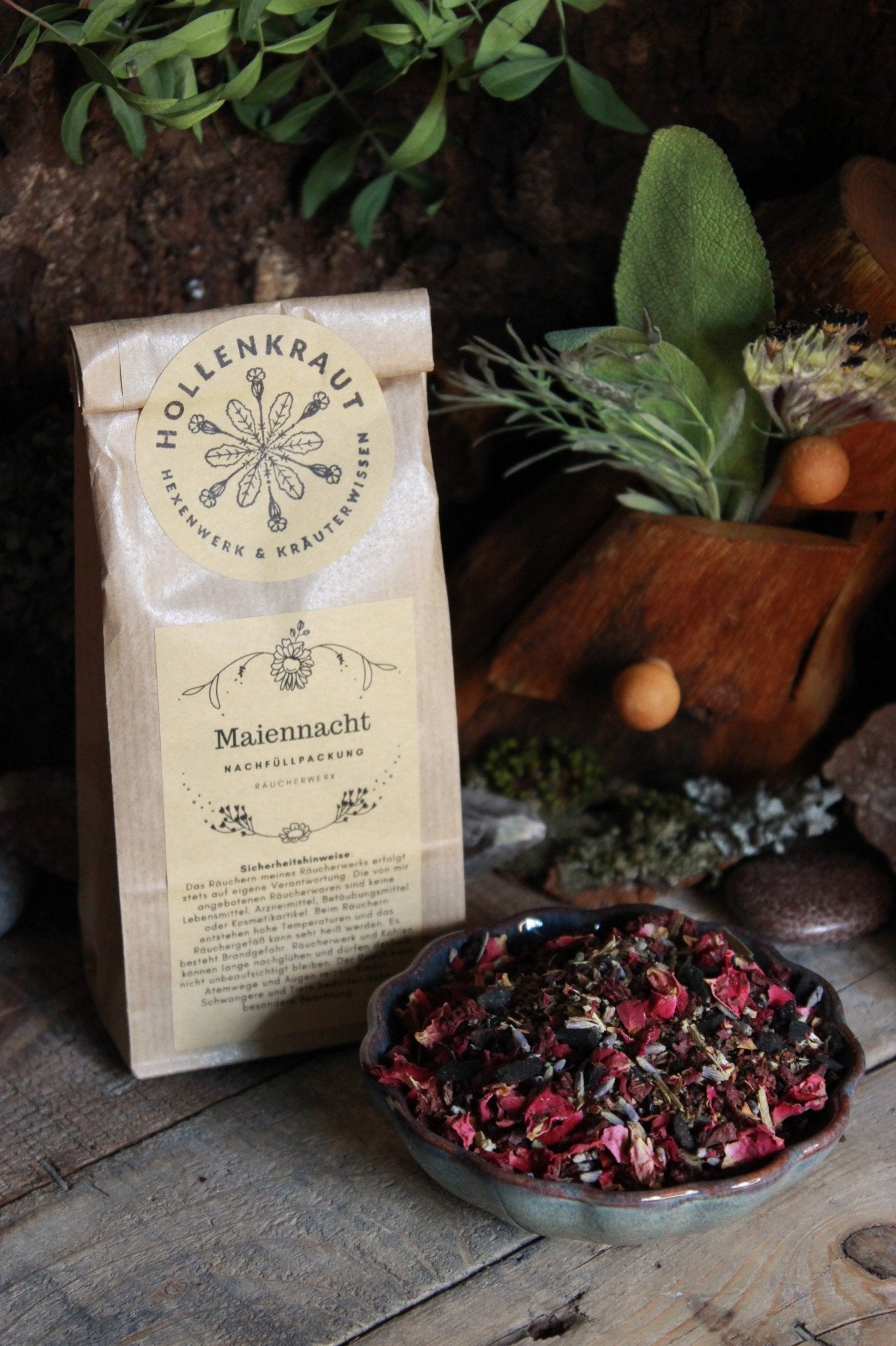 Eine Packung ‘Hollenkraut Hexenwerk & Kräuterwissen’ neben einer Schale mit getrockneten Blumen und Kräutern auf einem rustikalen Holztisch.