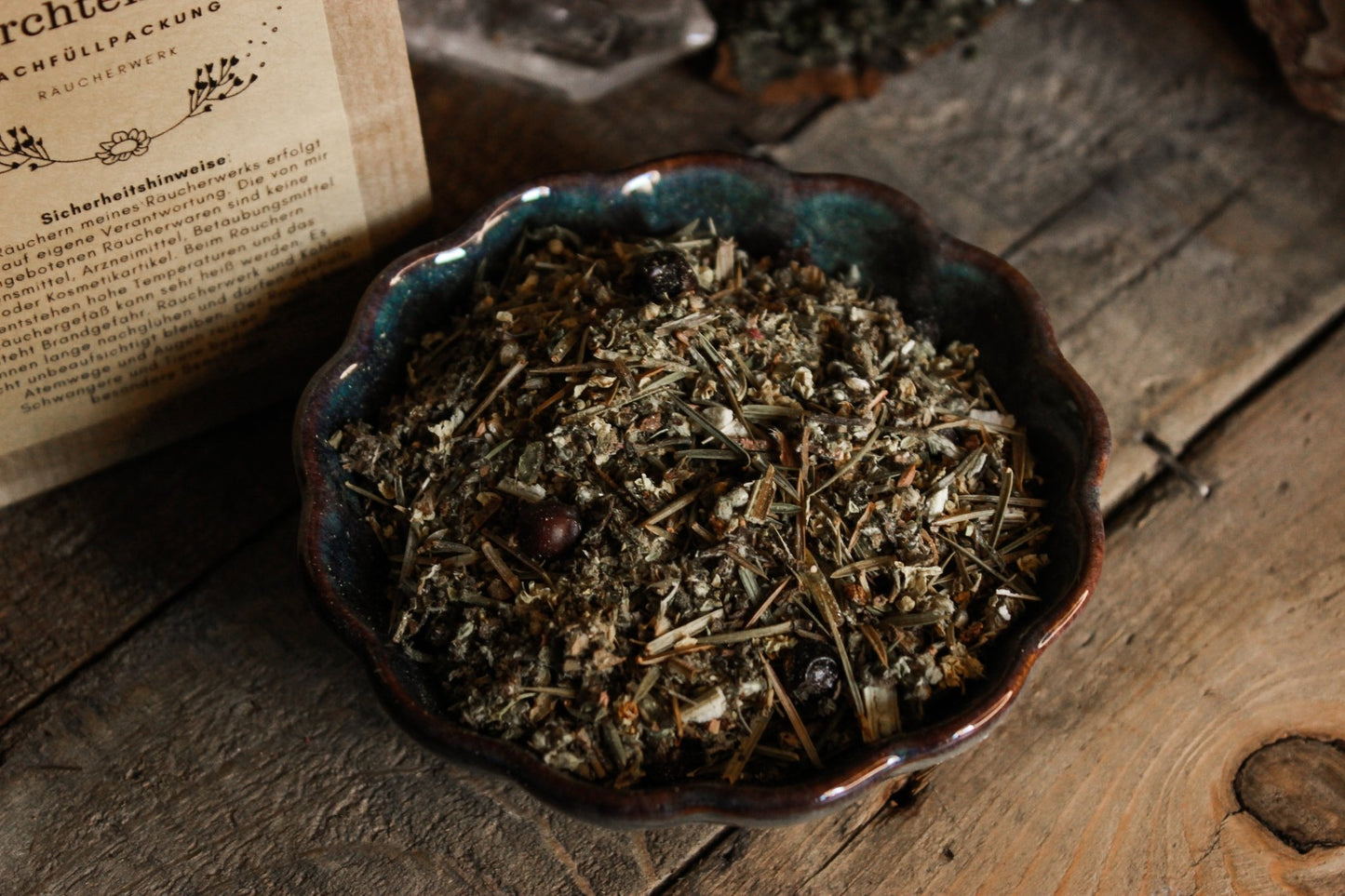 Räucherwerk Perchtenlauf - Räucherwerk -Das Bild zeigt eine Mischung aus getrockneten Kräutern in einer künstlerischen Schale, die auf einem rustikalen Holztisch platziert ist. Eine Packung mit gedruckten Sicherheitshinweisen und einem floralen Design ist teilweise links von der Schale sichtbar. Die Kräuter sind fein gemischt.