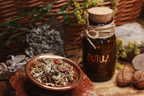 Räucherwerk Trotula Rustikale Darstellung einer dunklen Glasflasche mit der Aufschrift ‘TROTULA’, neben einer Kupferschale mit getrockneten Kräutern und Blumen, umgeben von Walnüssen und Steinen, mit einem Weidenkorb im Hintergrund.