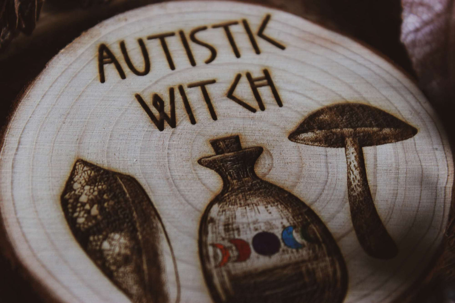 Nahaufnahme Wandbild aus Holz "Autistic Witch" - Handgefertigte Holzgravur mit dem Text ‘AUTISTIC WITCH’, umgeben von detaillierten Zeichnungen eines Blattes, einer Flasche mit farbiger Flüssigkeit und eines Pilzes.