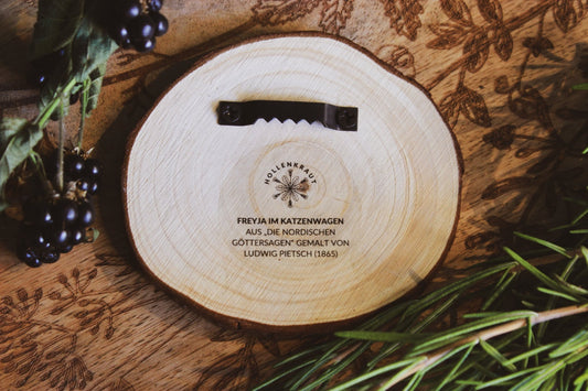 Wandbild aus Holz "Freyja im Katzenwagen" - Handgeschnitztes Holzkunstwerk mit graviertem Design und Text ‘FREYJA IM KATZENWAGEN AUS ,DIE NORDISCHEN GÖTTERSAGEN’ GEMALT VON LUDWIG PIETSCH (1865)', präsentiert auf einem rustikalen Holzhintergrund, dekoriert mit frischen Beeren und Blättern.