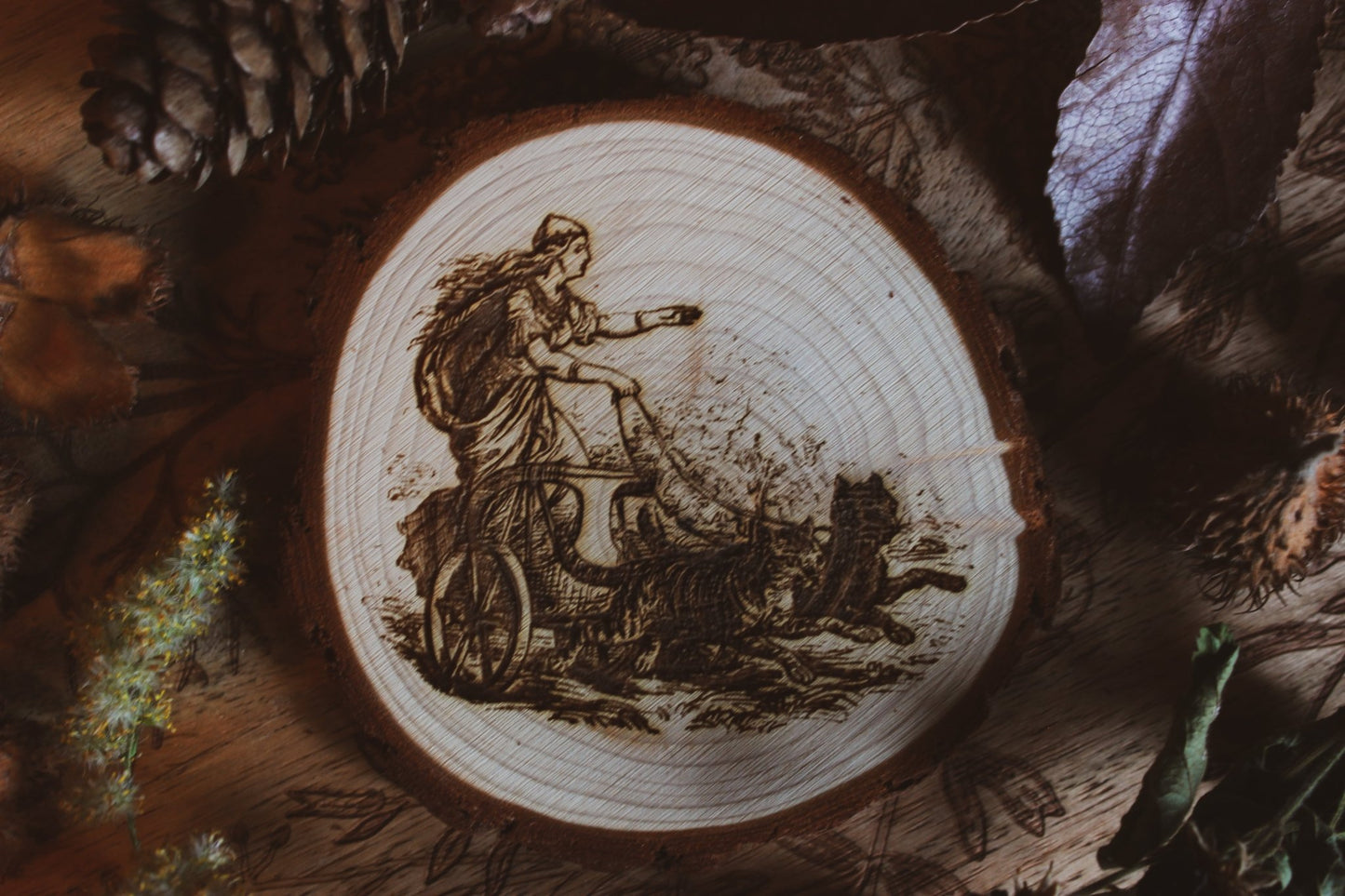 Wandbild aus Holz "Freyja im Katzenwagen" - “Handgefertigtes Kunstwerk auf einem kreisförmigen Holzstück, das eine Freya auf einem Zugwagen darstellt, Katzen ziehen ihn Das Kunstwerk ist umgeben von Naturmaterialien wie Tannenzapfen und Blättern.