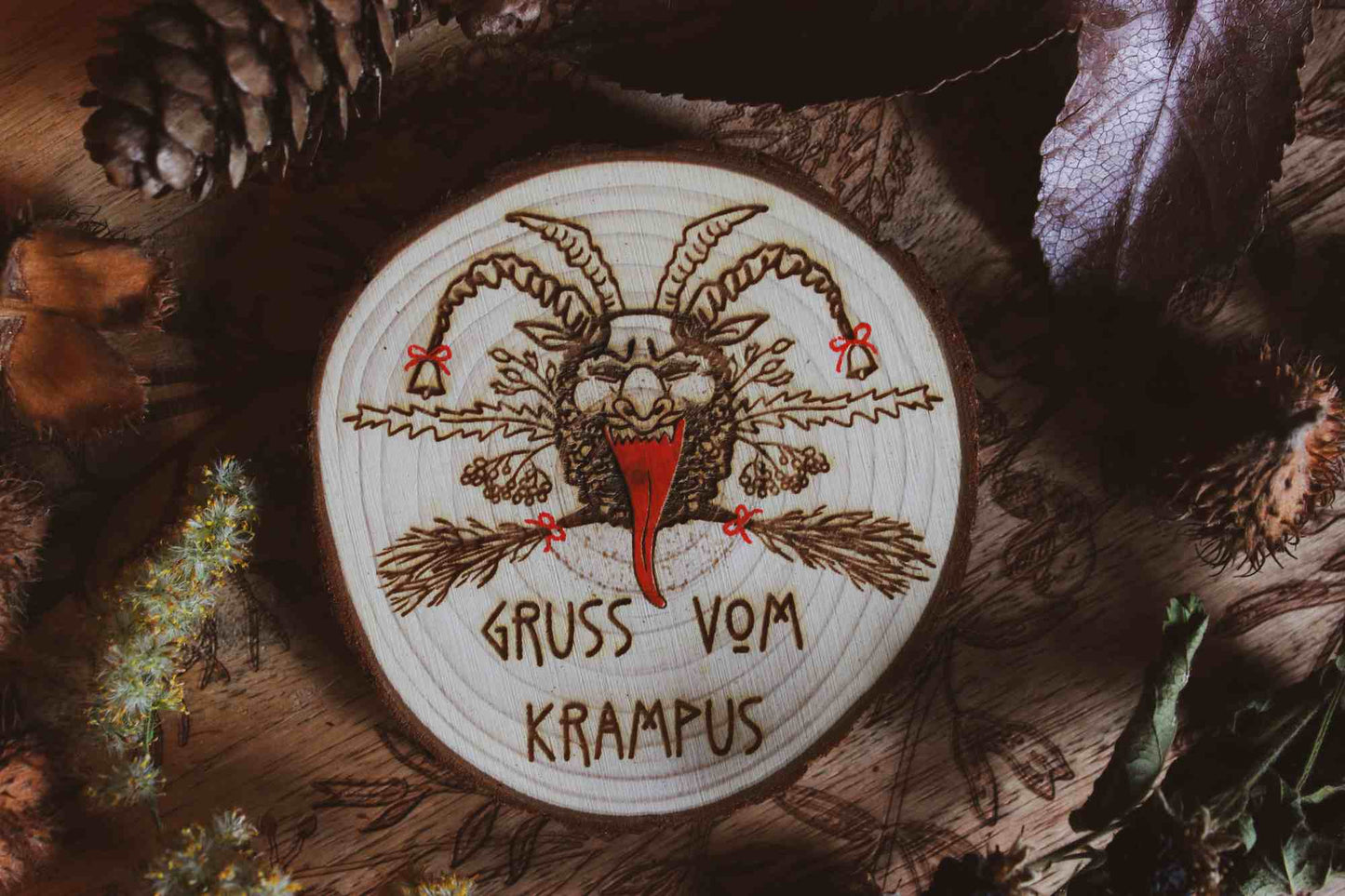 Wandbild aus Holz "Gruss vom Krampus" - Handgefertigtes Krampus-Kunstwerk auf einem runden Holzstück, umgeben von natürlichen Elementen wie Kiefernzapfen und getrockneten Blättern. Krampus ist mit Hörnern und einer langen Zunge dargestellt, was die typische Darstellung der Folklore-Kreatur widerspiegelt. Der Schriftzug ‘GRUSS VOM KRAMPUS’ ist am unteren Rand eingraviert. Die rustikale Erscheinung des Holzstücks und die schattige Beleuchtung verleihen dem Bild eine dunkle und stimmungsvolle Atmosphäre.