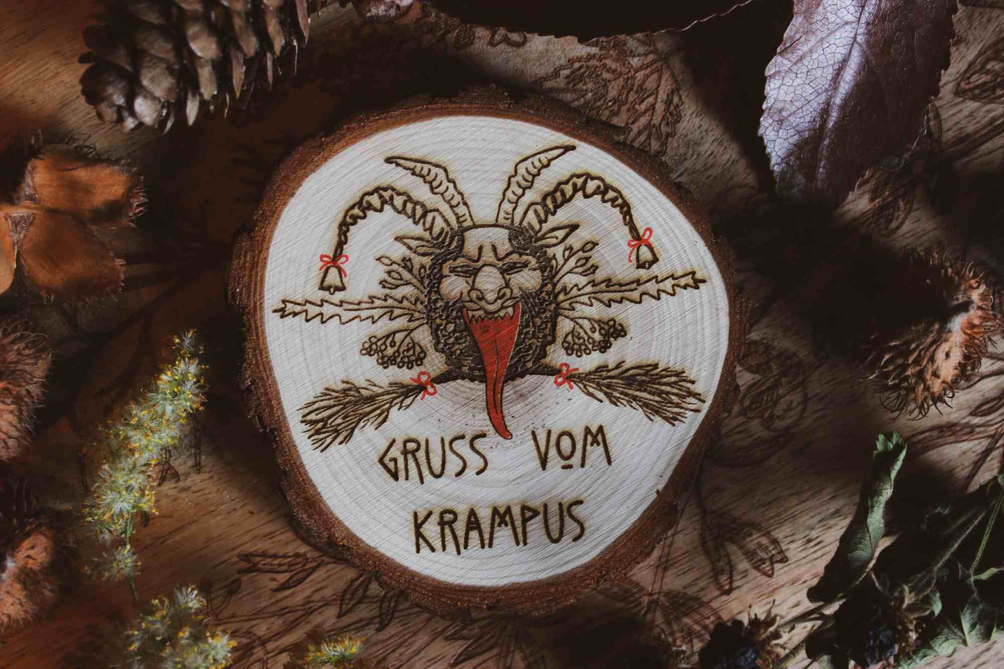Wandbild aus Holz "Gruss vom Krampus" -Handgefertigtes Krampus-Kunstwerk auf einem runden Holzstück, umgeben von natürlichen Elementen wie Kiefernzapfen und getrockneten Blättern. Krampus ist mit Hörnern und einer langen Zunge dargestellt, was die typische Darstellung der Folklore-Kreatur widerspiegelt. Der Schriftzug ‘GRUSS VOM KRAMPUS’ ist am unteren Rand eingraviert. Die rustikale Erscheinung des Holzstücks und die schattige Beleuchtung verleihen dem Bild eine dunkle und stimmungsvolle Atmosphäre.