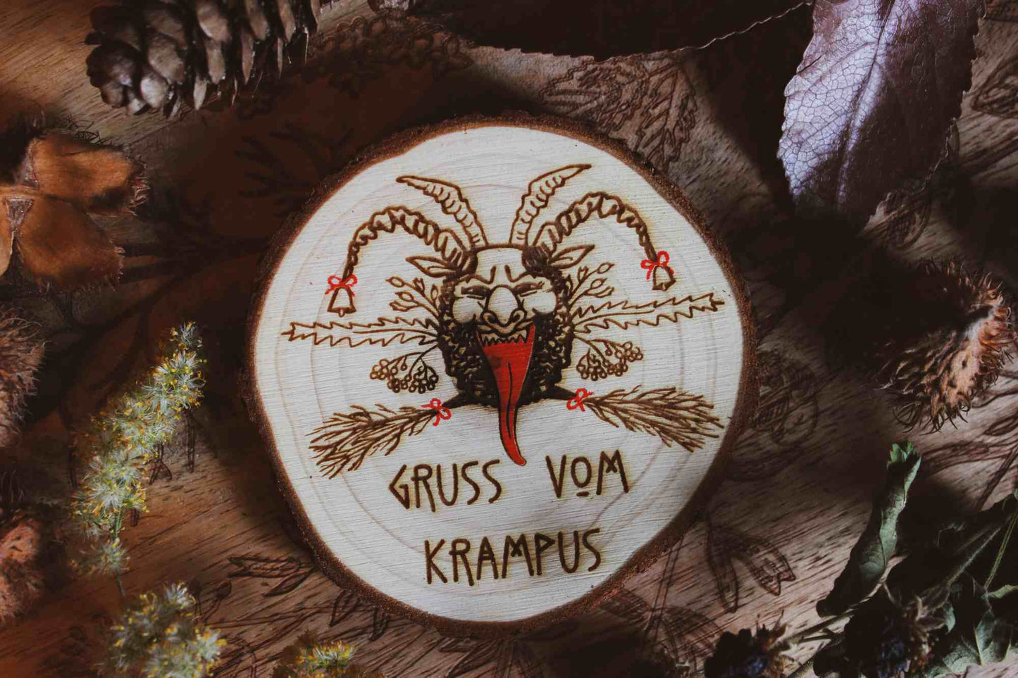 Wandbild aus Holz "Gruss vom Krampus" - Handgefertigtes Krampus-Kunstwerk auf einem runden Holzstück, umgeben von natürlichen Elementen wie Kiefernzapfen und getrockneten Blättern. Krampus ist mit Hörnern und einer langen Zunge dargestellt, was die typische Darstellung der Folklore-Kreatur widerspiegelt. Der Schriftzug ‘GRUSS VOM KRAMPUS’ ist am unteren Rand eingraviert. Die rustikale Erscheinung des Holzstücks und die schattige Beleuchtung verleihen dem Bild eine dunkle und stimmungsvolle Atmosphäre.
