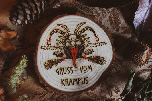Wandbild aus Holz "Gruss vom Krampus" -Handgefertigtes Krampus-Kunstwerk auf einem runden Holzstück, umgeben von natürlichen Elementen wie Kiefernzapfen und getrockneten Blättern. Krampus ist mit Hörnern und einer langen Zunge dargestellt, was die typische Darstellung der Folklore-Kreatur widerspiegelt. Der Schriftzug ‘GRUSS VOM KRAMPUS’ ist am unteren Rand eingraviert. Die rustikale Erscheinung des Holzstücks und die schattige Beleuchtung verleihen dem Bild eine dunkle und stimmungsvolle Atmosphäre.