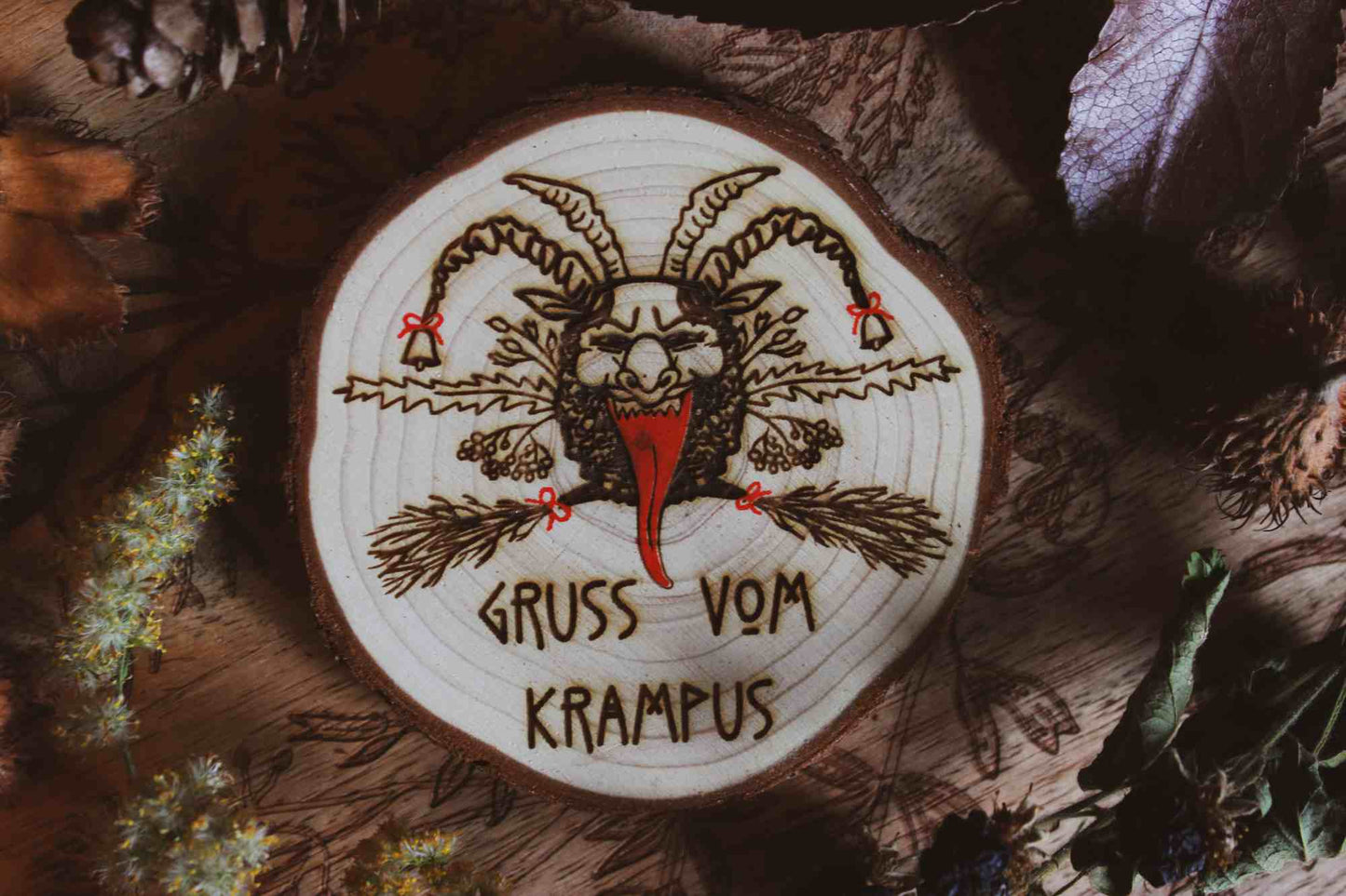 Wandbild aus Holz "Gruss vom Krampus" - Handgefertigtes Krampus-Kunstwerk auf einem runden Holzstück, umgeben von natürlichen Elementen wie Kiefernzapfen und getrockneten Blättern. Krampus ist mit Hörnern und einer langen Zunge dargestellt, was die typische Darstellung der Folklore-Kreatur widerspiegelt. Der Schriftzug ‘GRUSS VOM KRAMPUS’ ist am unteren Rand eingraviert. Die rustikale Erscheinung des Holzstücks und die schattige Beleuchtung verleihen dem Bild eine dunkle und stimmungsvolle Atmosphäre.