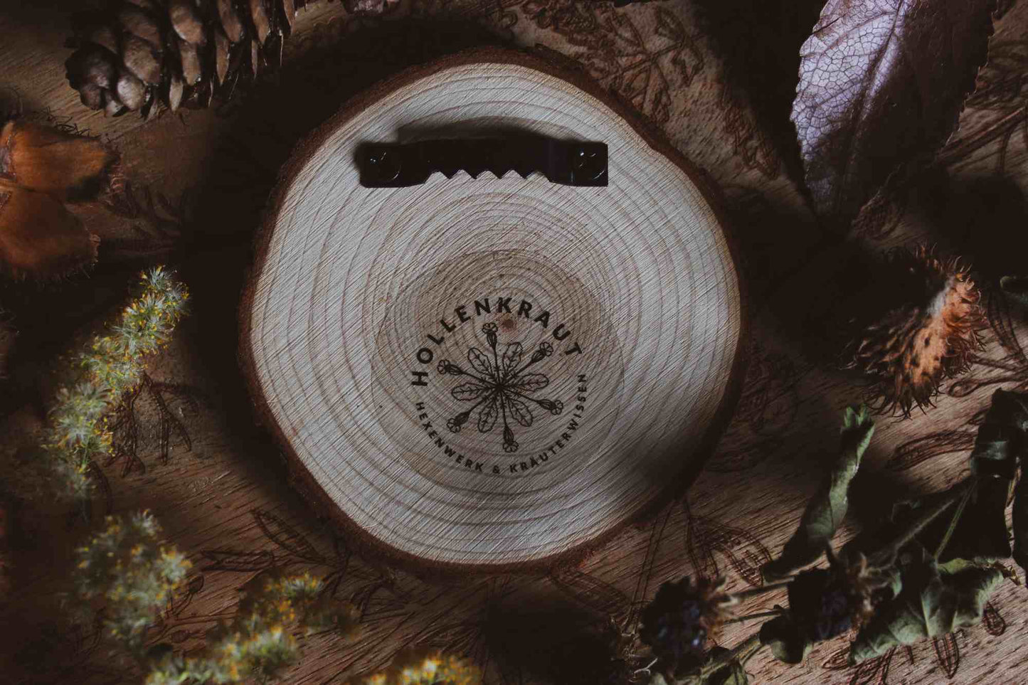 Rückseite Wandbild aus Holz "Gruss vom Krampus" -Rückseite mit Hollenkraut Logo und Aufhänger auf einem runden Holzstück, umgeben von natürlichen Elementen wie Kiefernzapfen und getrockneten Blättern. Krampus ist mit Hörnern und einer langen Zunge dargestellt, was die typische Darstellung der Folklore-Kreatur widerspiegelt. Die rustikale Erscheinung des Holzstücks und die schattige Beleuchtung verleihen dem Bild eine dunkle und stimmungsvolle Atmosphäre.