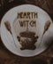 Wandbild aus Holz "Hearth Witch" -Bild eines hölzernen Gegenstands mit der Inschrift ‘Hearth Witch’, auf der Scheibe sieht man 2 Besen die einen Hexenkessel umrahmen umgeben von natürlichen Elementen wie Tannenzapfen und Blumen,