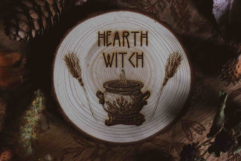 Wandbild aus Holz "Hearth Witch" -Bild eines hölzernen Gegenstands mit der Inschrift ‘Hearth Witch’, auf der Scheibe sieht man 2 Besen die einen Hexenkessel umrahmen umgeben von natürlichen Elementen wie Tannenzapfen und Blumen,