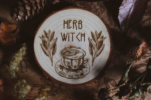 Wandbild aus Holz "Herb Witch" - Wandbild aus Holz "Herb Witch" - Dieses Bild zeigt eine kunstvoll gravierte Holzscheibe mit der Inschrift “HERB WITCH”, umgeben von einer natürlichen und mystischen Atmosphäre. Die Gravur stellt eine Tasse Tee dar, die von zwei Zweigen mit Blättern flankiert wird,