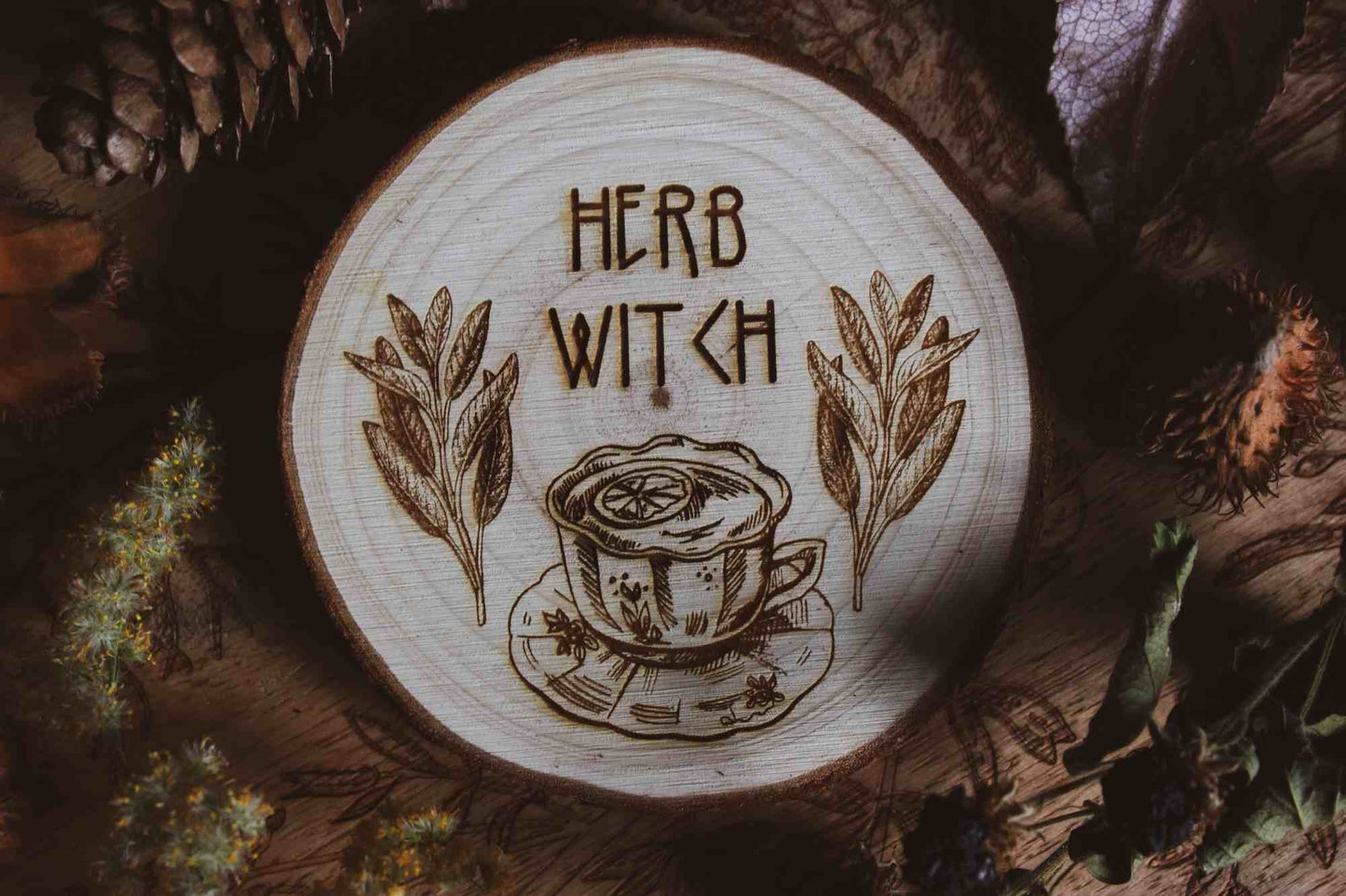 Wandbild aus Holz "Herb Witch" - Dieses Bild zeigt eine kunstvoll gravierte Holzscheibe mit der Inschrift “HERB WITCH”, umgeben von einer natürlichen und mystischen Atmosphäre. Die Gravur stellt eine Tasse Tee dar, die von zwei Zweigen mit Blättern flankiert wird,