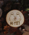 Wandbild aus Holz "No Feet" - Handgefertigtes Holzstück mit einer eingravierten Illustration von zwei Geistern und dem Text ‘NO FEET’, umgeben von natürlichen Elementen wie Tannenzapfen, Steinen und einer Froschstatue.