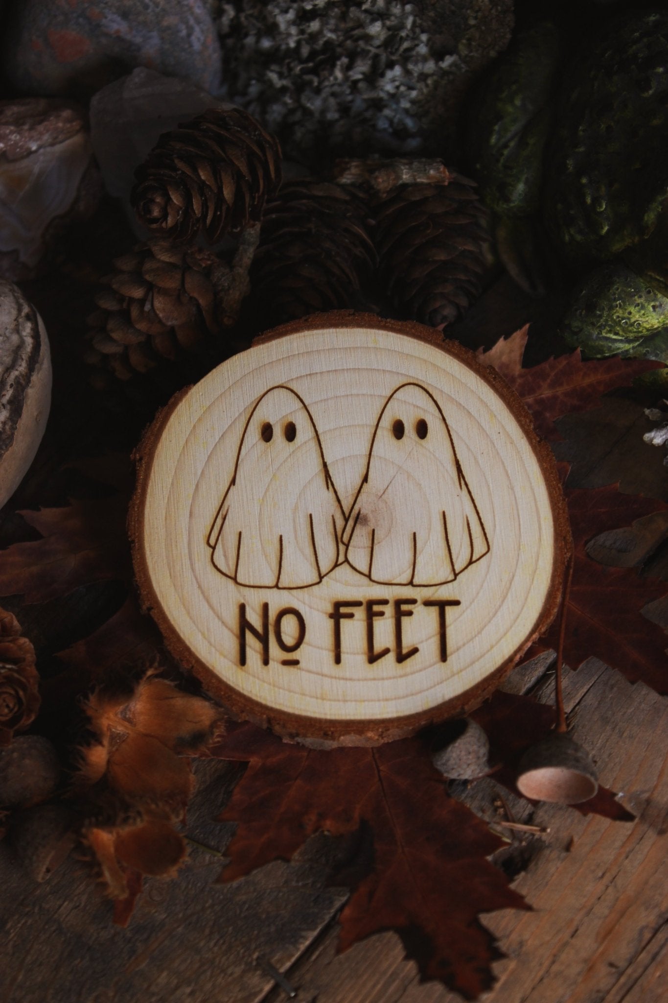 Wandbild aus Holz "No Feet" - Handgefertigtes Holzstück mit einer eingravierten Illustration von zwei Geistern und dem Text ‘NO FEET’, umgeben von natürlichen Elementen wie Tannenzapfen, Steinen und einer Froschstatue.