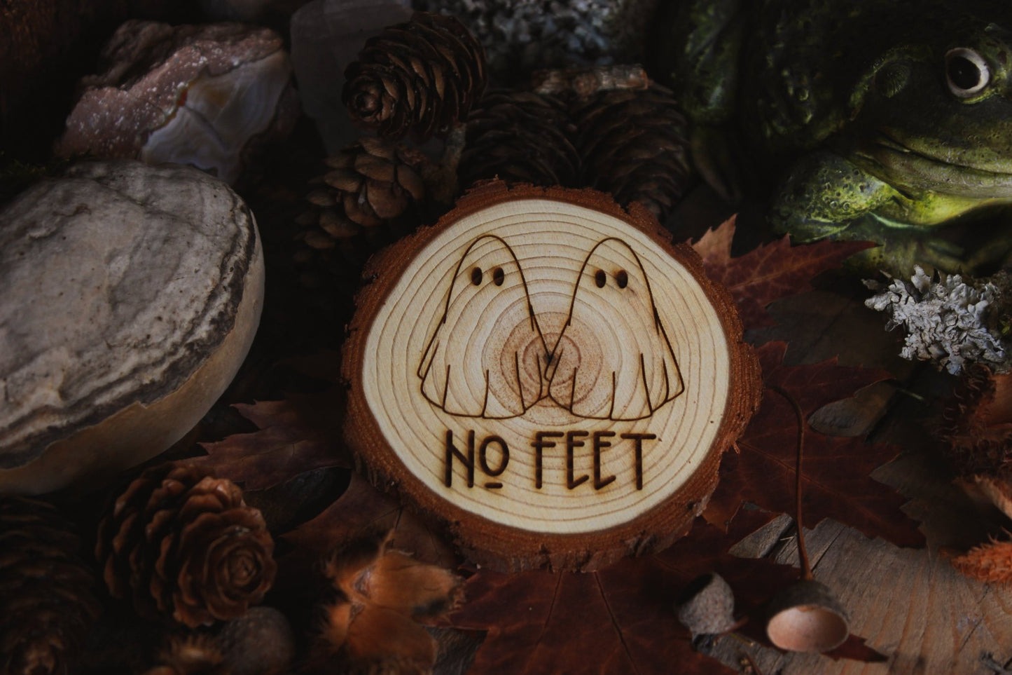 Wandbild aus Holz "No Feet" - Handgefertigtes Holzstück mit einer eingravierten Illustration von zwei Geistern und dem Text ‘NO FEET’, umgeben von natürlichen Elementen wie Tannenzapfen, Steinen und einer Froschstatue.