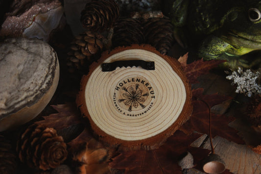 Rückseite Wandbild aus Holz "No Feet" - Handgefertigtes Holzstück mit dem Hollenkraut Logo und einer Aufhängung, umgeben von natürlichen Elementen wie Tannenzapfen, Steinen und einer Froschstatue.