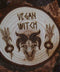 Wandbild aus Holz "Vegan Witch" - Wandbild aus Holz "Vegan Witch" - Das Bild zeigt ein handgefertigtes Kunstwerk mit der Inschrift “VEGAN WITCH”, umgeben von einer dunklen, mystischen Atmosphäre und natürlichen Elementen wie Tannenzapfen und Blättern.