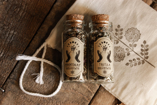 Zwei Glasflaschen ‘Perchtenlauf’ und ‘Trotula’, gefüllt mit getrockneten Kräutern, liegen auf einem rustikalen Holztisch neben einem Stück Stoff mit Blumenmuster.