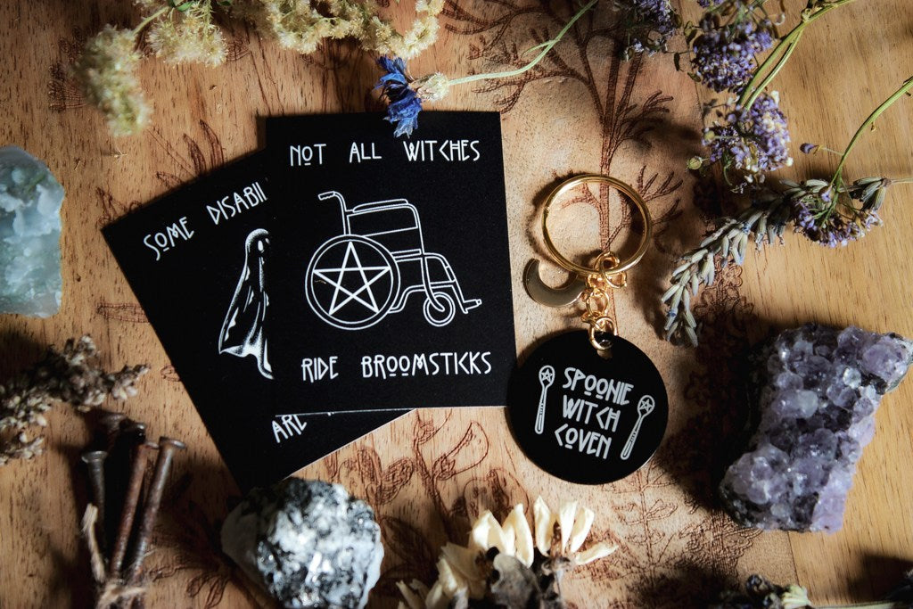 Zwei schwarze Sticker und ein Schlüsselanhänger in Schwarz und Gold. Auf der schwarzen Scheibe steht in weiß: Spoonie Witch Coven. Auf dem obersten vollständig sichtbaren Sticker steht: Not all witches ride broomsticks.