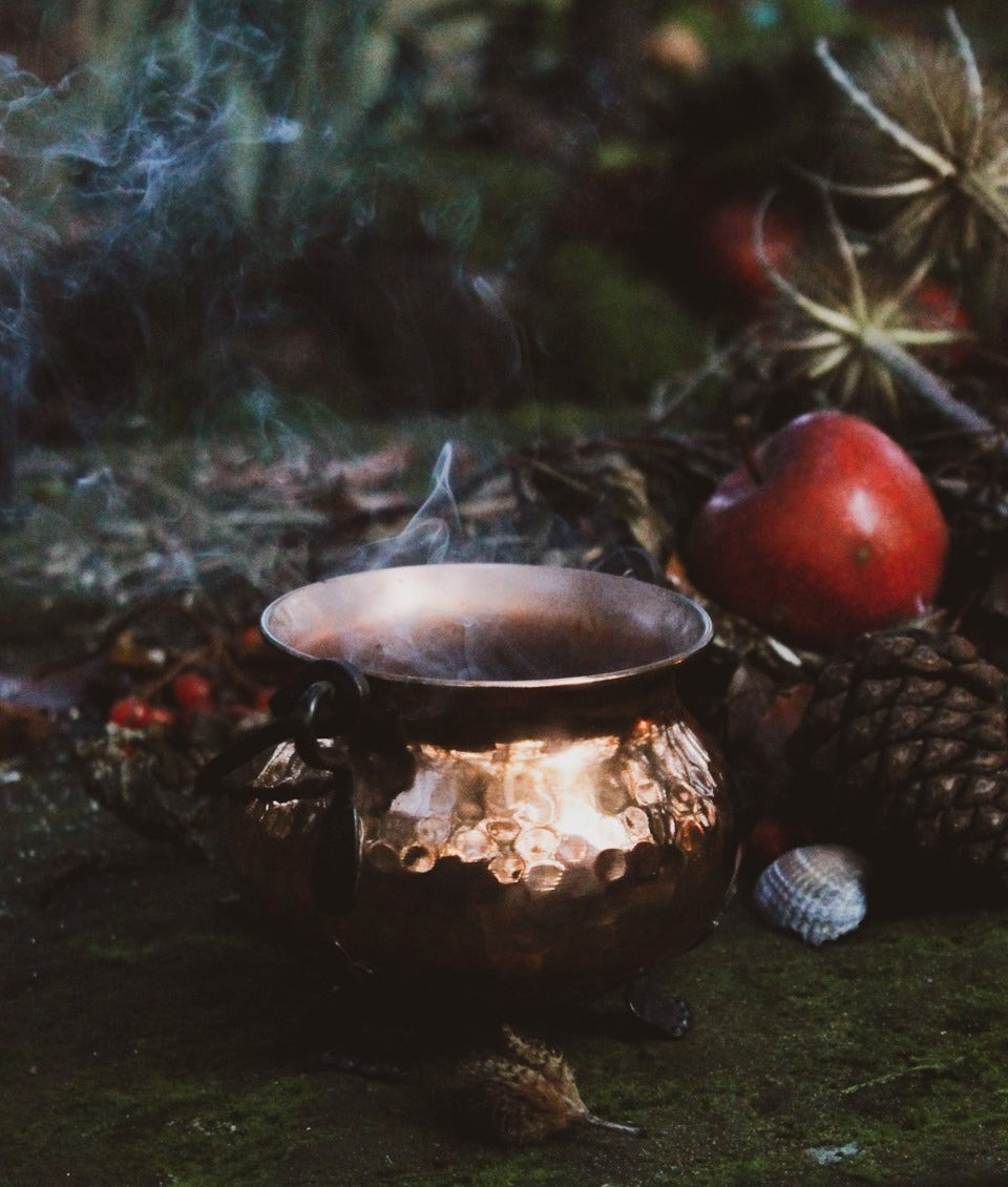 Ein Kupferkessel aus dem Rauch aufsteigt. Daneben liegen verschiedene Naturmaterialien wie ein Apfel, Muscheln, Bucheckern.