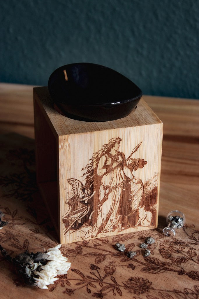 Auf einem Holztisch steht eine Duftlampe mit Holzsockel und schwarzen Keramik-Einsätzen. Auf das Holz ist ein Bild der Göttin Freya mit ihrem Katzen eingebrannt.