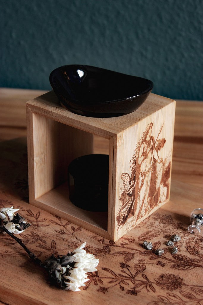 Auf einem Holztisch steht eine Duftlampe mit Holzsockel und schwarzen Keramik-Einsätzen. Auf das Holz ist ein Bild der Göttin Freya mit ihrem Katzen eingebrannt.