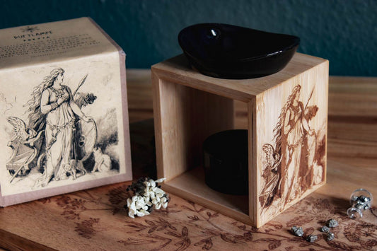 Auf einem Holztisch steht eine Duftlampe mit Holzsockel und schwarzen Keramik-Einsätzen. Auf das Holz ist ein Bild der Göttin Freya mit ihrem Katzen eingebrannt.
