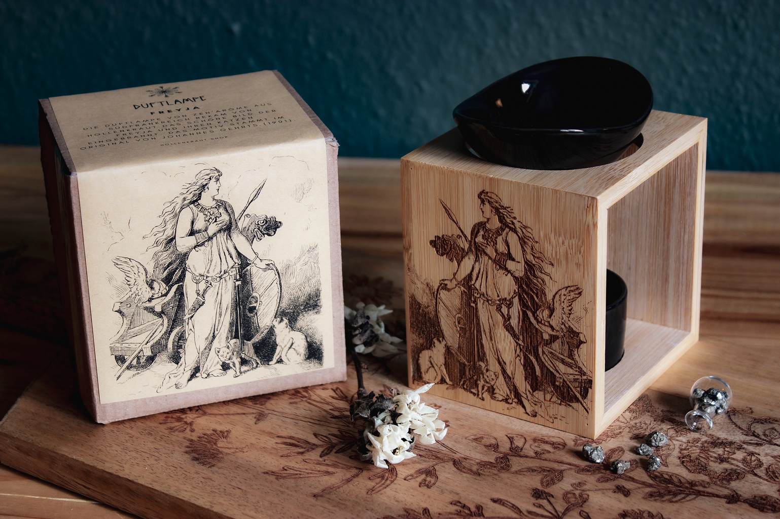 Auf einem Holztisch steht eine Duftlampe mit Holzsockel und schwarzen Keramik-Einsätzen. Auf das Holz ist ein Bild der Göttin Freya mit ihrem Katzen eingebrannt. Daneben steht ein Produktkarton der das gleiche Bildnis aufgedruckt hat.