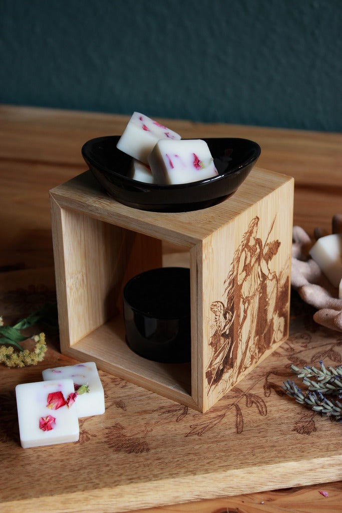 Auf einem Holztisch steht eine Duftlampe mit Holzsockel und schwarzen Keramik-Einsätzen. Auf das Holz ist ein Bild der Göttin Freya mit ihrem Katzen eingebrannt. Oben in der Schale liegt weißes Duftwachs mit Rosenblüten darin.