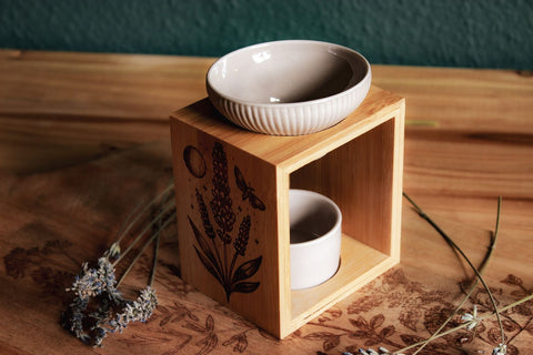 Eine Duftlampe aus Holz und grau-beiger Keramik. In das Holz eingebrannt ist das Motiv eines Lavendels, daneben der Mond, eine Motte und kleine Sterne.